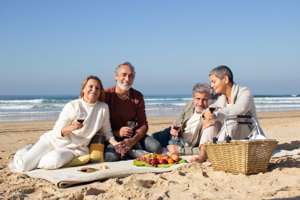 two-lovely-senior-couples-enjoying-picnic-sandy-beach-old-friends-hanging-out-together-drinking-wine-recalling-good-times-people-smiling-looking-camera-leisure-friendship-concept